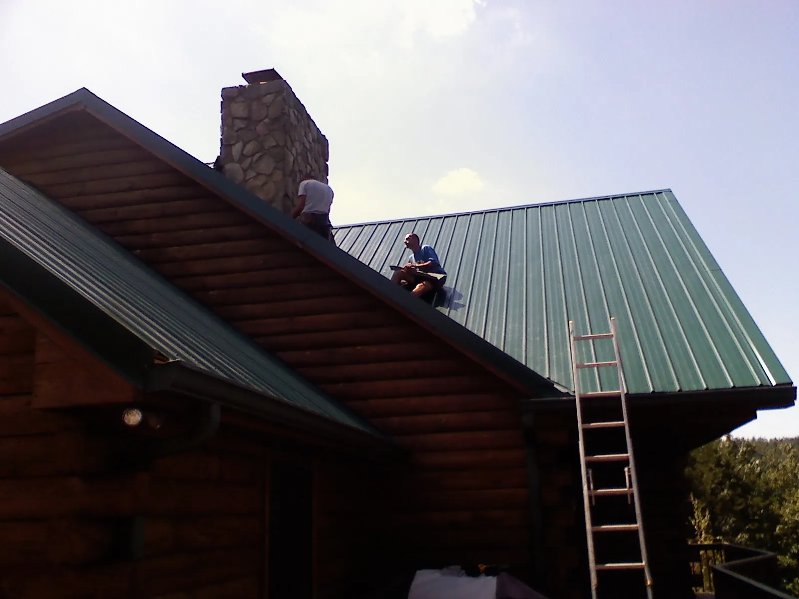 Roofers installing green metal panels beside a stone chimney on a log home in Washburn